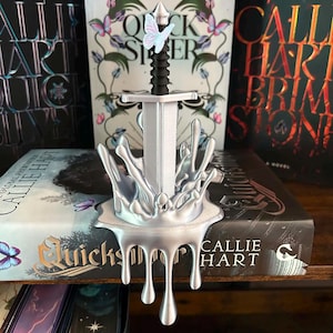 May include: A silver-colored sword sculpture featuring a black handle and a small butterfly accent. The sword is set within a base that resembles a metallic liquid splash, with drips extending downward from the front edge of the base.