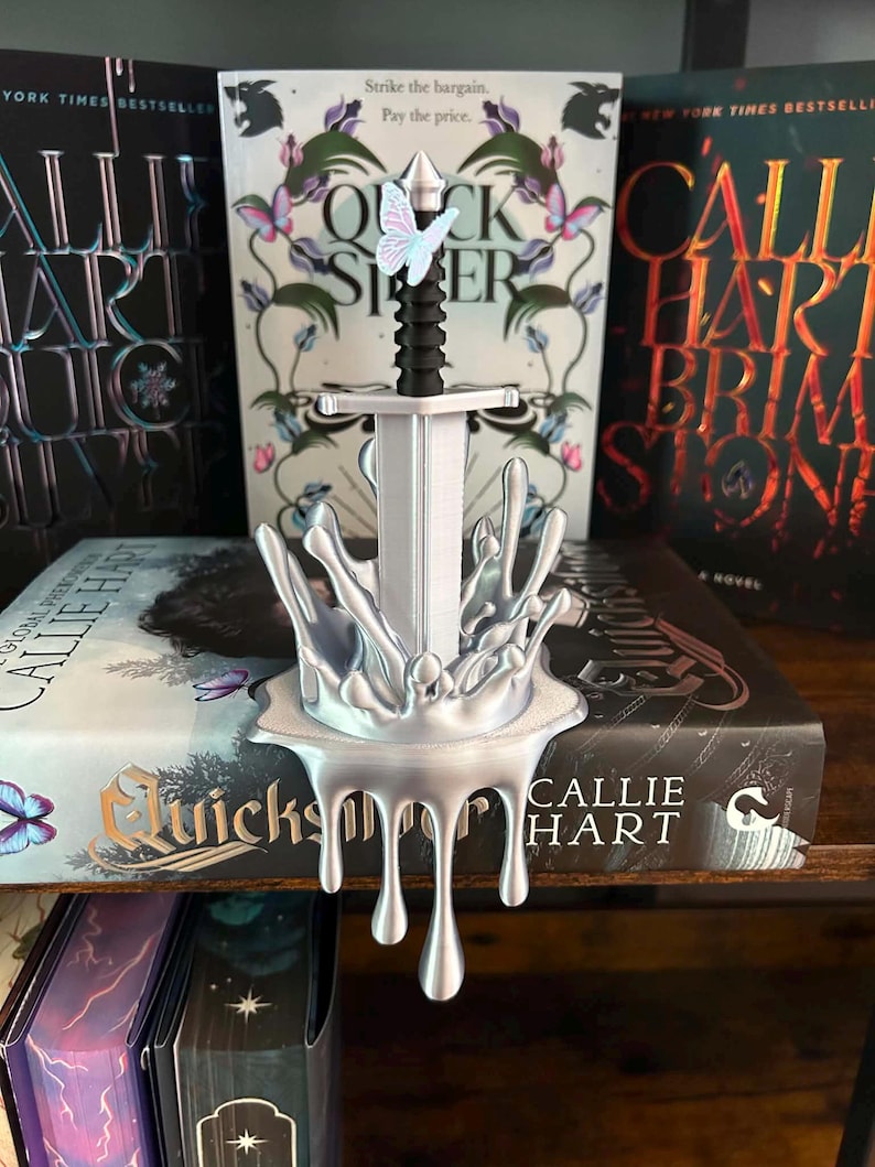 May include: A silver-colored sword sculpture featuring a black handle and a small butterfly accent. The sword is set within a base that resembles a metallic liquid splash, with drips extending downward from the front edge of the base.