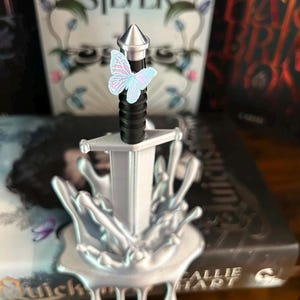 May include: A silver-colored sword with a black handle and a pointed pommel, standing upright in a silver-colored base that resembles a liquid splash. A small, light blue and pink butterfly is attached to the upper part of the sword handle.