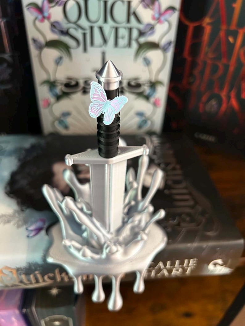 May include: A silver-colored sword with a black handle and a pointed pommel, standing upright in a silver-colored base that resembles a liquid splash. A small, light blue and pink butterfly is attached to the upper part of the sword handle.