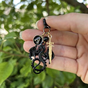 May include: A black dragon charm with gold accents, a sword charm, and a black and gold circular charm. The dragon has outstretched wings and a curled tail. The charms are attached to a black clasp.