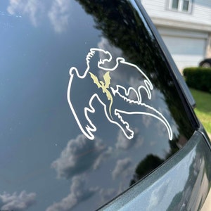 May include: A white silhouette of a dragon with a smaller dragon inside, on a black car window.