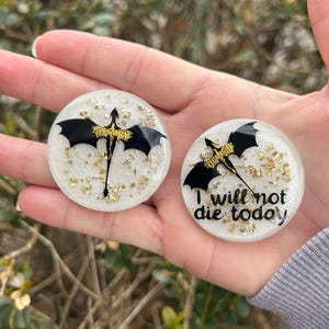 May include: Two white round resin phone grips with gold glitter. One grip features a black silhouette of a dragon with gold accents. The other grip features a black silhouette of a dragon with gold accents and the text "I will not die today".