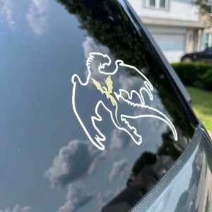 May include: A white vinyl sticker of a dragon silhouette with a gold outline on a black car window.