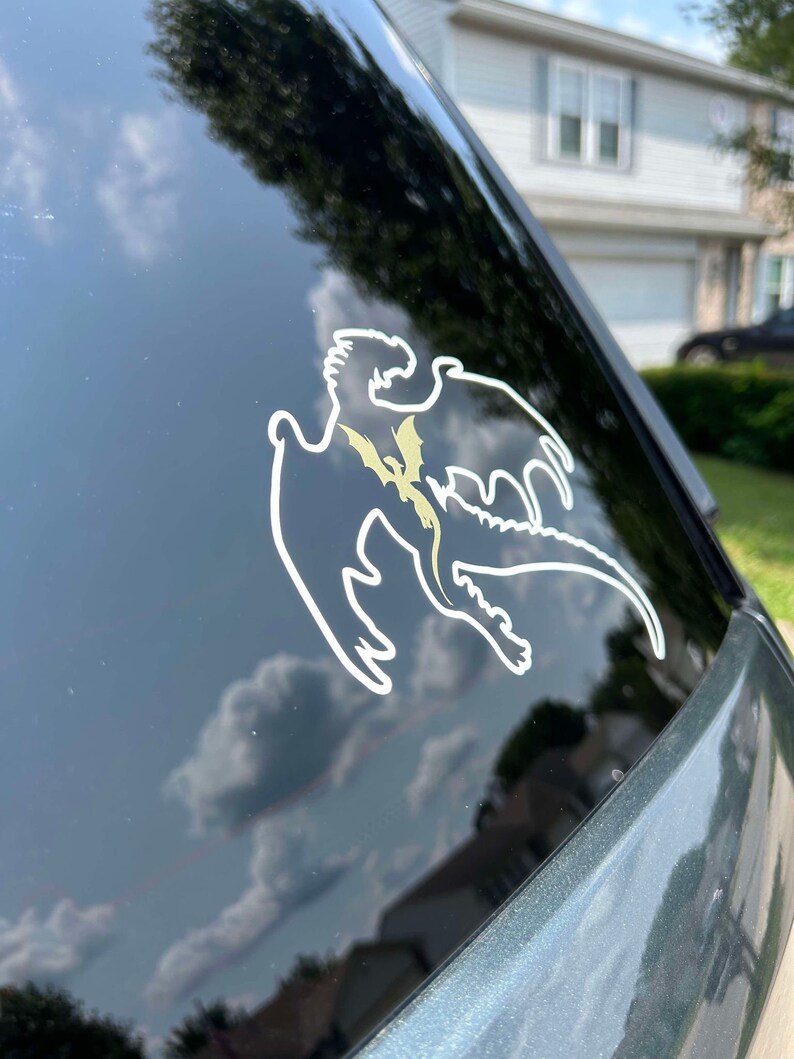 May include: A white vinyl sticker of a dragon silhouette with a gold outline on a black car window.