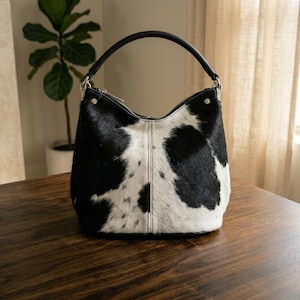 K&ouml;nnte beinhalten: Eine Handtasche im Hobo-Stil mit einem schwarz-wei&szlig;en Kuhfellmuster und einer texturierten Oberfl&auml;che aus echtem Haar. Die Tasche verf&uuml;gt &uuml;ber einen einzelnen schwarzen Ledergriff mit silberfarbenen Metallbeschl&auml;gen und einen Rei&szlig;verschluss.