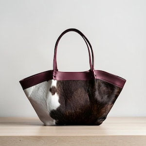 May include: A tote bag with a cowhide pattern in white and brown, complemented by a burgundy leather trim and handles. The bag is set on a light-colored wooden surface, showcasing its design and texture.