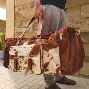 Puede incluir: Una bolsa de viaje grande confeccionada en piel de vaca marr&oacute;n y blanca. La bolsa cuenta con un bolsillo exterior prominente con dos correas de hebilla, asas de cuero resistentes y un compartimento principal con cremallera y herrajes dorados.