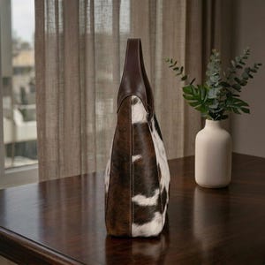May include: A tall tote bag with a brown leather handle and trim. The body of the bag is made of cowhide with a brown and white spotted pattern. The bag is standing on a dark brown wooden surface.