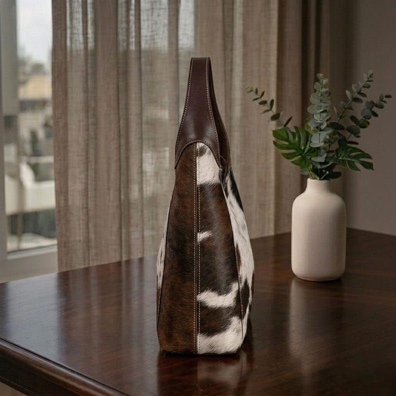 May include: A tall tote bag with a brown leather handle and trim. The body of the bag is made of cowhide with a brown and white spotted pattern. The bag is standing on a dark brown wooden surface.