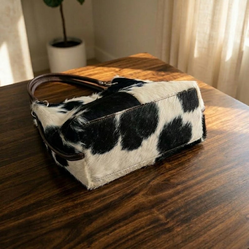 May include: A rectangular handbag with a cowhide pattern in black and white. The bag has a brown leather handle and a structured shape. The bag is sitting on a wooden surface.