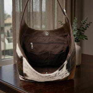 May include: A brown and white cowhide hobo bag with a leather strap and interior pockets. The bag has a magnetic snap closure and a zippered pocket. The interior is a dark brown fabric. The bag is sitting on a dark wooden surface.