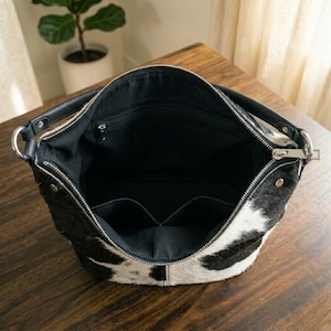 K&ouml;nnte beinhalten: Eine schwarz-wei&szlig;e Hobo-Tasche aus Kuhfell mit schwarzem Innenfutter. Die Tasche verf&uuml;gt &uuml;ber einen Rei&szlig;verschluss oben, zwei schwarze Ledergriffe mit silberfarbenen Metallbeschl&auml;gen sowie eine Innentasche mit Rei&szlig;verschluss und zwei Einsteckf&auml;chern.