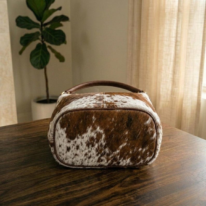 May include: A brown and white cowhide handbag with a rounded shape and a dark brown handle. The bag is sitting on a dark wooden surface. The bag has a unique spotted pattern.