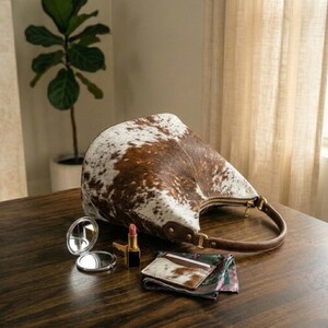 May include: A brown and white cowhide leather handbag with a zipper closure and a brown leather handle. A small silver compact mirror, lipstick, and a cowhide leather cardholder are on a wooden surface.