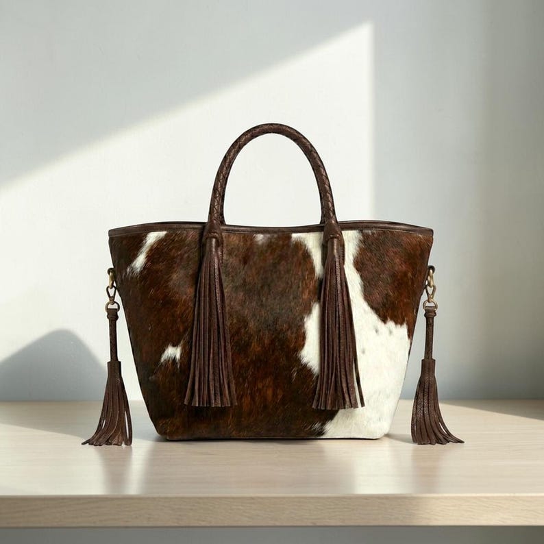 May include: A brown and white cowhide tote bag with a braided handle and brown leather trim. The bag features two long brown tassels and a detachable shoulder strap. The bag is sitting on a light brown surface.