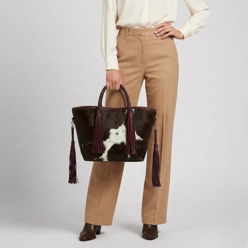 May include: A cowhide tote bag in brown and white, featuring a braided handle and burgundy tassels. The bag has a dark brown trim and a distinctive white and brown spotted pattern. The bag is held by a person wearing tan trousers and a white shirt.