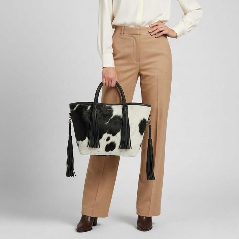 May include: A black and white cowhide tote bag with black leather handles and trim. The bag features long black tassels on the sides. The person holding the bag is wearing tan trousers and a white shirt.