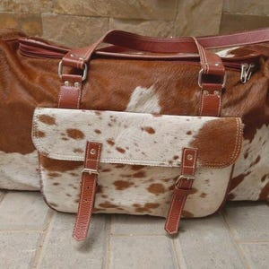Puede incluir: Una bolsa de viaje de piel de vaca marr&oacute;n y blanca con un acabado texturizado. La bolsa incluye un bolsillo frontal con dos correas de cuero con hebillas, herrajes de metal plateado, dos asas superiores y compartimentos laterales con cremallera.