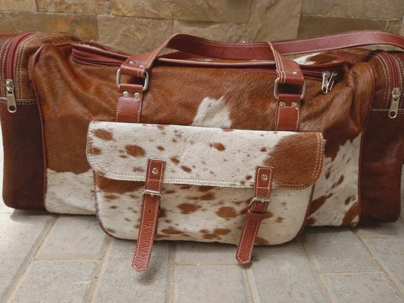 Puede incluir: Una bolsa de viaje de piel de vaca marr&oacute;n y blanca con un acabado texturizado. La bolsa incluye un bolsillo frontal con dos correas de cuero con hebillas, herrajes de metal plateado, dos asas superiores y compartimentos laterales con cremallera.