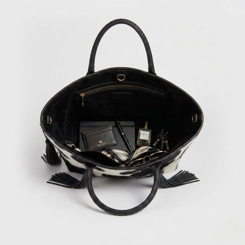 May include: Black and white cowhide patterned tote bag with black handles and tassels. The bag contains a wallet, pen, notebook, perfume, keys, and sunglasses. Internal measurements.