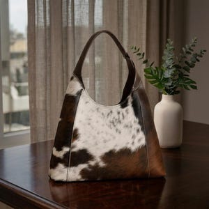 May include: A cowhide leather shoulder bag with a brown leather handle. The bag features a spotted pattern of brown and white, with brown leather sides and a curved shape. The bag is sitting on a dark wooden table.