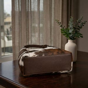 May include: A cowhide shoulder bag with a brown leather base and handle. The bag features a pattern of brown and white patches. The bag is sitting on a dark wooden table.