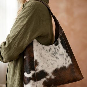 May include: A brown, white, and black cowhide shoulder bag with a dark brown leather strap. The bag has a unique pattern and a curved top. The bag is carried over the shoulder.