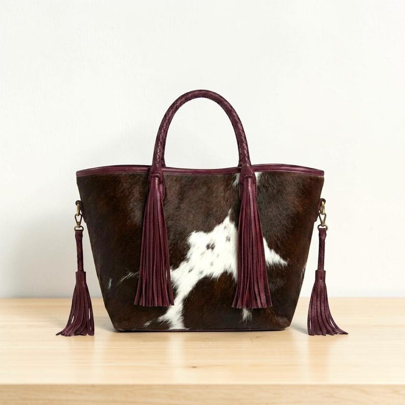 May include: A brown and white cowhide tote bag with a braided handle and burgundy leather trim. The bag features long burgundy tassels and a detachable shoulder strap. The bag is sitting on a light brown wooden surface.