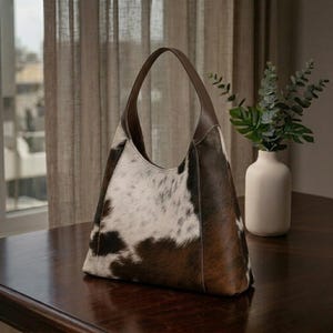 May include: A brown and white cowhide leather shoulder bag with a brown leather handle. The bag is on a dark wooden table. A white vase with green plants is in the background.