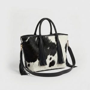 May include: A black and white cowhide tote bag with a braided handle and a detachable shoulder strap. The bag features black tassels and gold-coloured hardware. The bag is approximately 30 cm wide and 25 cm tall.