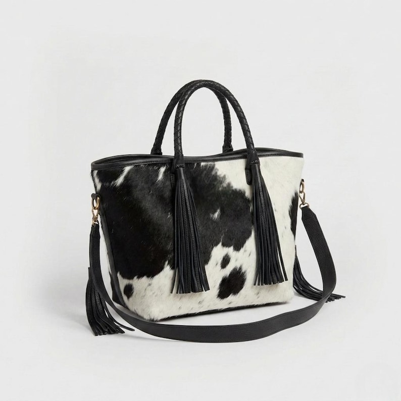 May include: A black and white cowhide tote bag with a braided handle and a detachable shoulder strap. The bag features black tassels and gold-coloured hardware. The bag is approximately 30 cm wide and 25 cm tall.