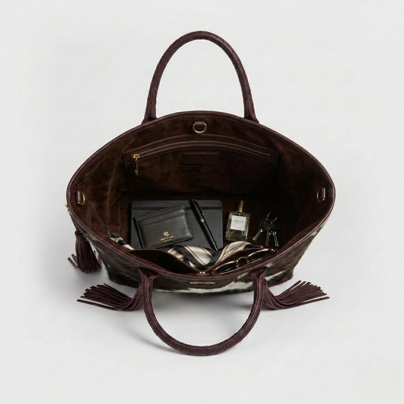 May include: A large open tote bag with a dark brown exterior and a lighter brown interior. The bag has a cowhide pattern with white and brown patches. Inside are a wallet, pen, notebook, perfume, keys, and a scarf.