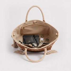 May include: Open tote bag with a cowhide pattern in shades of brown and cream. The interior is beige and contains a wallet, pen, notebook, perfume, keys, and a striped scarf. The bag has two handles and decorative tassels.
