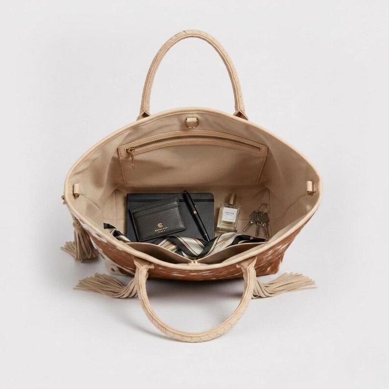 May include: Open tote bag with a cowhide pattern in shades of brown and cream. The interior is beige and contains a wallet, pen, notebook, perfume, keys, and a striped scarf. The bag has two handles and decorative tassels.