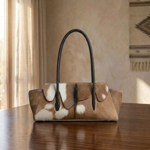 May include: A structured handbag featuring a cowhide exterior with a natural brown, white, and black spotted pattern. The bag has a horizontal rectangular shape with a flap closure and two tall, thin, black leather handles attached to the top.