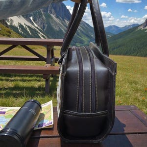 May include: A dark brown leather briefcase with double zippers and a handle is held up against a scenic mountain backdrop. A black water bottle and a map are on a wooden picnic table. The bag has a structured design.