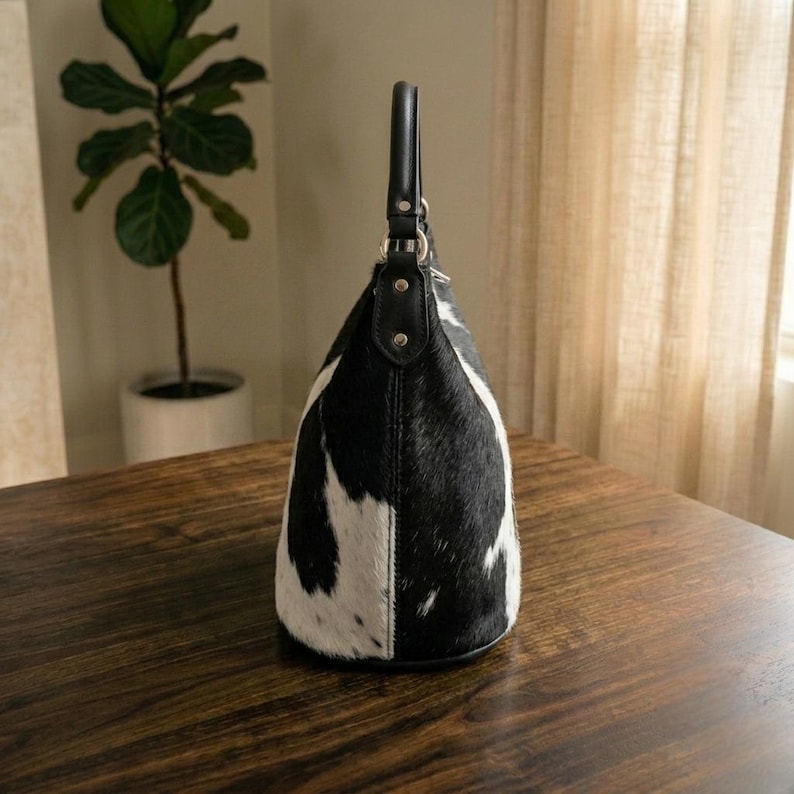 K&ouml;nnte beinhalten: Eine Handtasche im Hobo-Stil mit schwarz-wei&szlig;em Kuhfellmuster und strukturierter Felloberfl&auml;che. Die Tasche verf&uuml;gt &uuml;ber einen einzelnen schwarzen Ledergriff, der mit silberfarbenen Metallbeschl&auml;gen und Nieten befestigt ist und eine geschwungene Form aufweist.