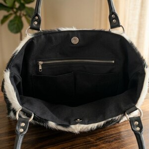 May include: A black and white faux fur tote bag with black leather handles and silver metal hardware. The interior is black with a zippered pocket and two open pockets. The bag is open, revealing the interior.