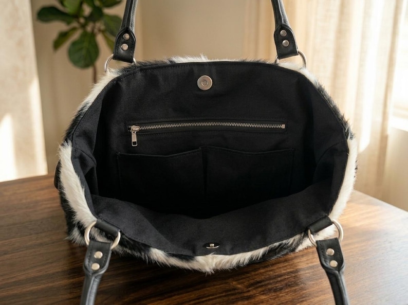 May include: A black and white faux fur tote bag with black leather handles and silver metal hardware. The interior is black with a zippered pocket and two open pockets. The bag is open, revealing the interior.