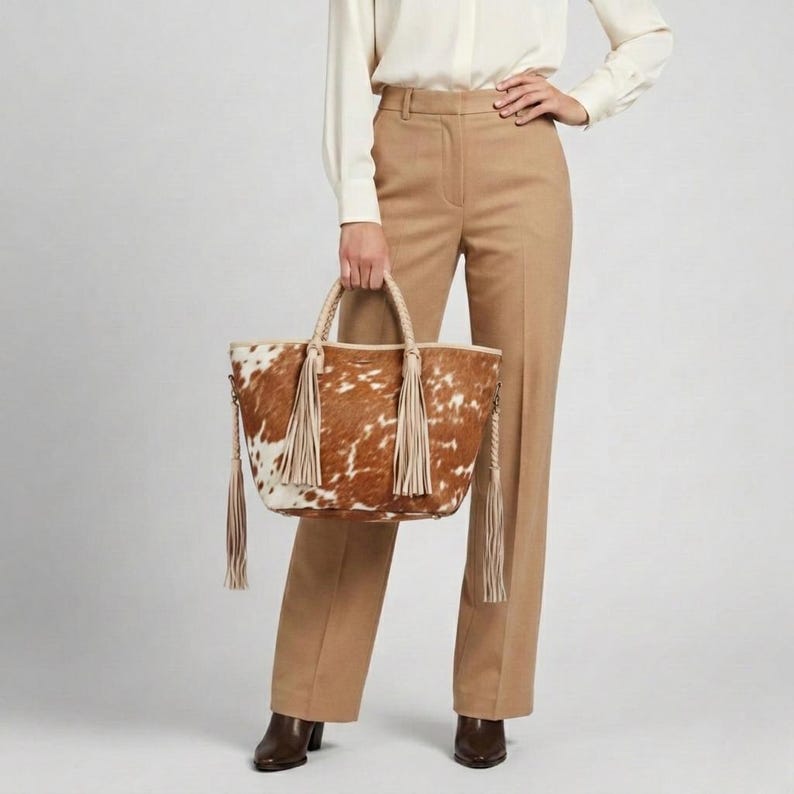 May include: A brown and white cowhide tote bag with braided handles and long cream-coloured tassels. The bag is held by a person wearing tan trousers and a white blouse. The bag's design features a natural pattern.