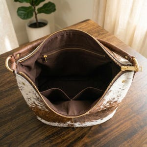 May include: Open view of a cowhide shoulder bag with a brown leather handle and gold-colored zipper. The bag's interior is brown with multiple pockets. The exterior features a mix of brown and white cowhide.