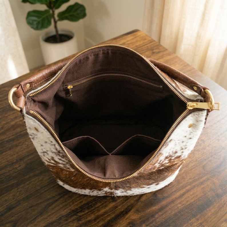 May include: Open view of a cowhide shoulder bag with a brown leather handle and gold-colored zipper. The bag's interior is brown with multiple pockets. The exterior features a mix of brown and white cowhide.