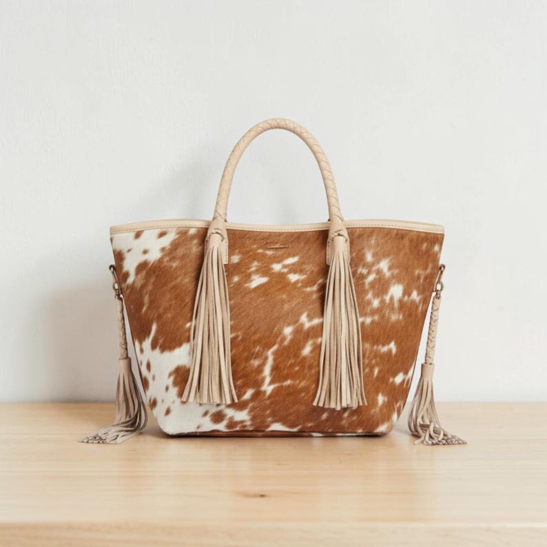 May include: A brown and white cowhide tote bag with a braided handle and long beige tassels. The bag has a light beige trim and a detachable shoulder strap. The bag is sitting on a wooden surface.
