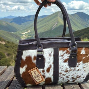 May include: A brown and white cowhide leather handbag with dark brown trim and handles. The bag has a rectangular shape with a zipper closure and a tag that reads "Genuine Leather". The bag is held up against a scenic mountain backdrop.