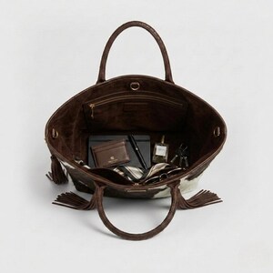 May include: A brown and white cowhide tote bag with brown leather handles. Inside, there's a wallet, pen, notebook, perfume, and keys. The bag has a suede interior and tassels on the sides. The bag's dimensions are approximately 30 cm wide.