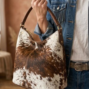 May include: A brown and white cowhide shoulder bag with a curved shape. The bag has a brown leather handle and trim, with a gold zipper closure. The bag is being held by a person wearing a denim jacket.