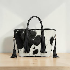 May include: A black and white cowhide-patterned tote bag. It features a black braided handle and black leather tassels. The bag is set on a wooden surface, showcasing its design and texture.
