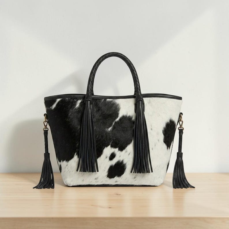 May include: A black and white cowhide-patterned tote bag. It features a black braided handle and black leather tassels. The bag is set on a wooden surface, showcasing its design and texture.