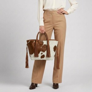 May include: A cowhide tote bag with a brown and white spotted pattern. The bag has brown leather handles and tassels. The person is wearing tan trousers and a white blouse.